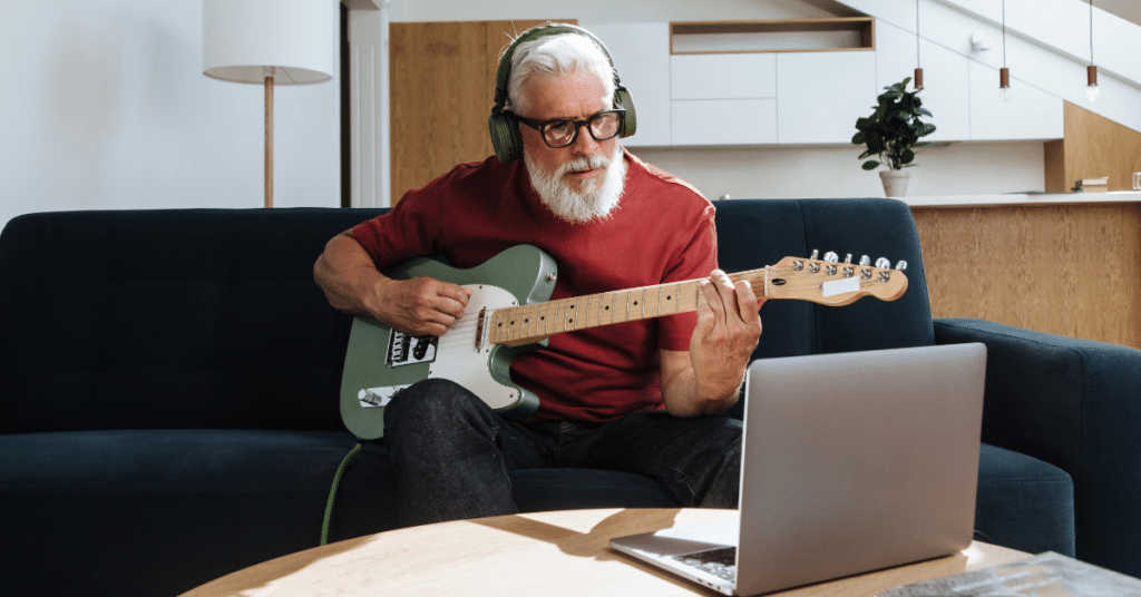 Mann mit E-Gitarre und Kopfhörern lernt online im Wohnzimmer vor dem Laptop – Rock & Pop Gitarrenkurs der New Music Academy.